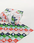 Single sheet of Winter Checkerboard gift wrap. Pink, blue, red, green, and yellow squares with neon icons of UFOs, hearts with eyes, moths, moons with stars, Saturn, shooting stars, moon phases, lightning bolts, moths, Hamsa hands, and a brightly burning star.