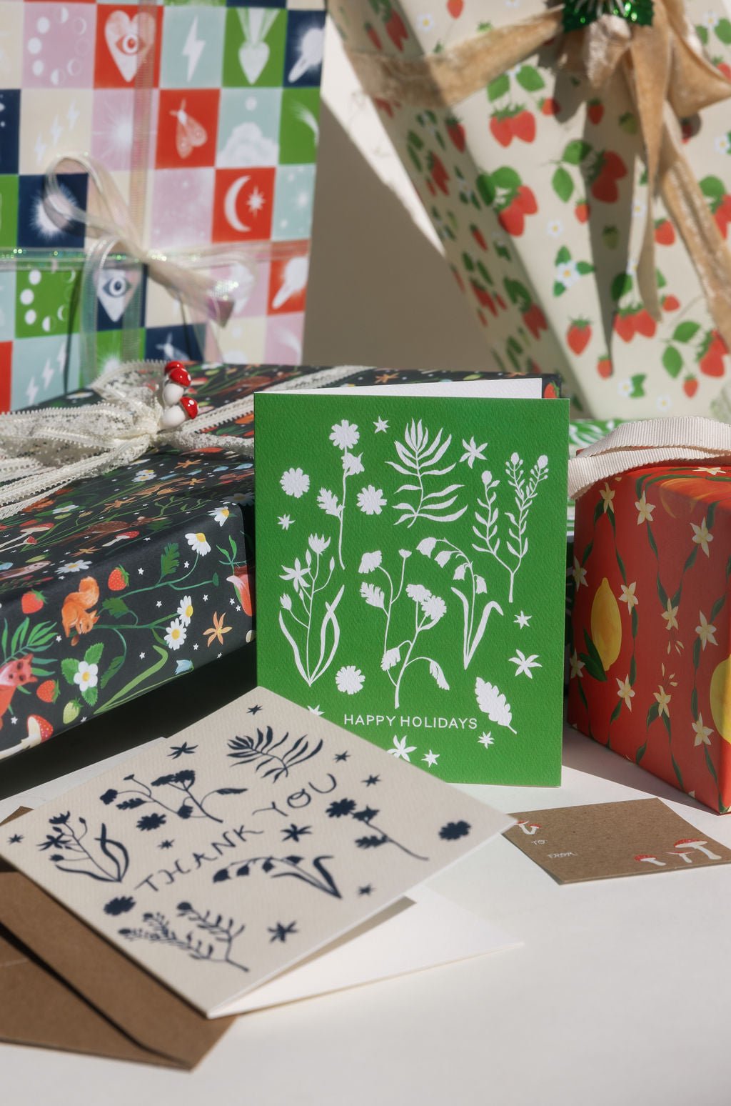 Our new forest flowers design of white outlines of forest flowers on a primary green background with the words "Happy Holidays" in caps lock text at the bottom.