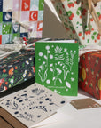 Our new forest flowers design of white outlines of forest flowers on a primary green background with the words "Happy Holidays" in caps lock text at the bottom.