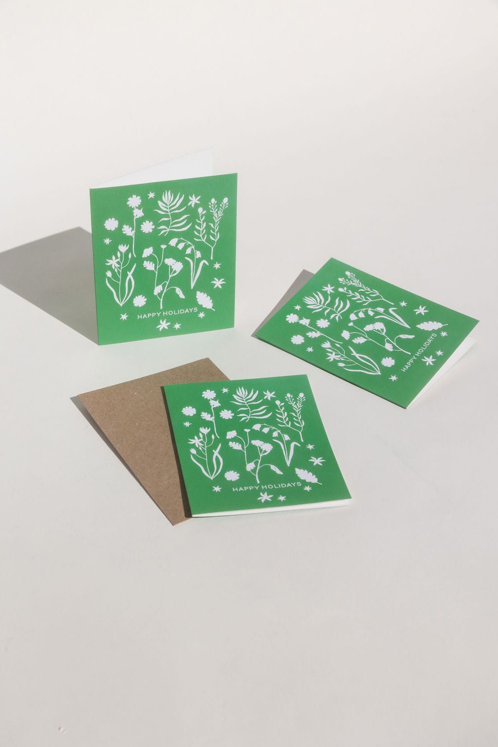 Our new forest flowers design of white outlines of forest flowers on a primary green background with the words "Happy Holidays" in caps lock text at the bottom.