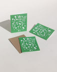 Our new forest flowers design of white outlines of forest flowers on a primary green background with the words "Happy Holidays" in caps lock text at the bottom.