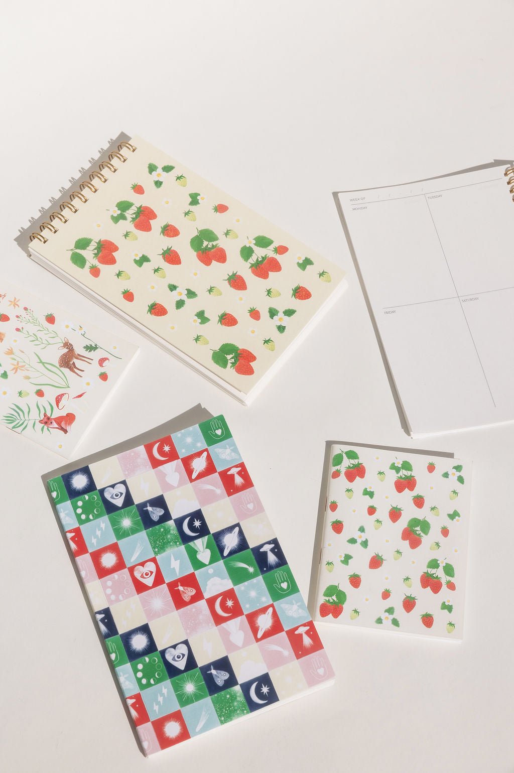 Holiday Checkerboard journal. Pink, blue, red, green, and yellow squares with neon icons of UFOs, hearts with eyes, moths, moons with stars, Saturn, shooting stars, moon phases, lightning bolts, moths, Hamsa hands, and a brightly burning star. Shown with our Strawberries & Cream taskpad, Strawberries pocket notebook, and planner.