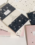 Adelfi notebook trio on a white background. One notebook has a cream background with black stars and comets, the second is navy blue with mystical outer space icons, and the third is pink with stars and eclipsing moons.