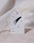 A single Adelfi letterpressed gift tag with a comet and stars and the words "to" and "from" in black ink on cream card stock leaning against a white fern on a gray background.