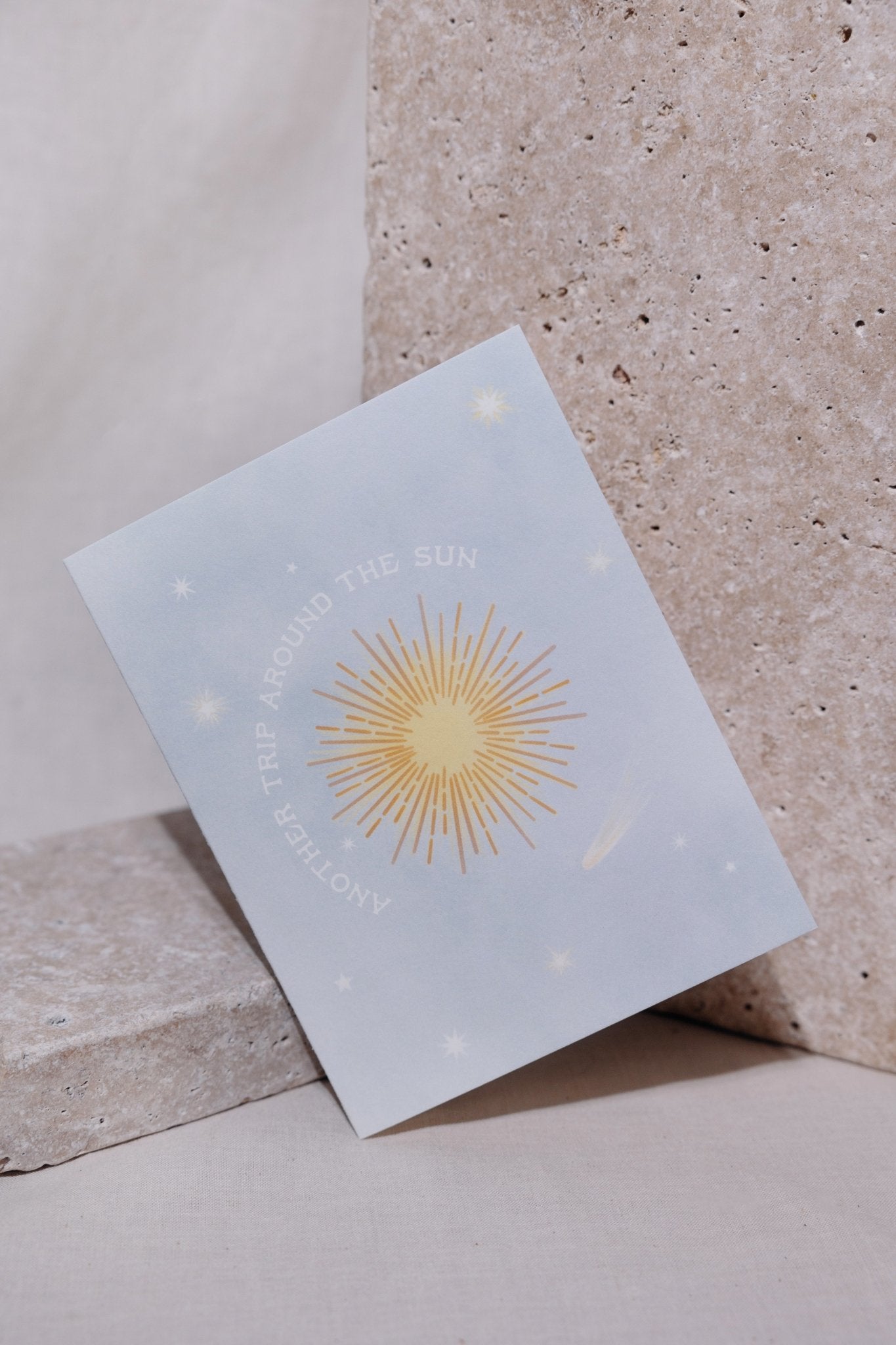 Greeting card with a place blue background, stars, and the words &quot;Another Trip Around the Sun&quot; circling around a yellow sun. Card is propped up on a beige background and is leaning at an angle against a light gray stone.