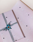 A present wrapped in gift wrap with a pale pink background, scattered with eclipsed moons and shooting stars, and a silver bow, placed on top of a single sheet of the same gift wrap.