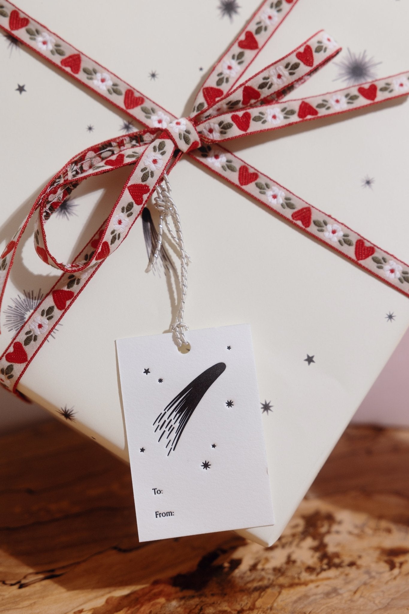 An Adelfi letterpressed gift tag with black comets, stars, and the words "to" and "from" in black ink on cream card stock attached by silver string to a present wrapped in cream and black comets Adelfi gift wrap with red hearts ribbon.