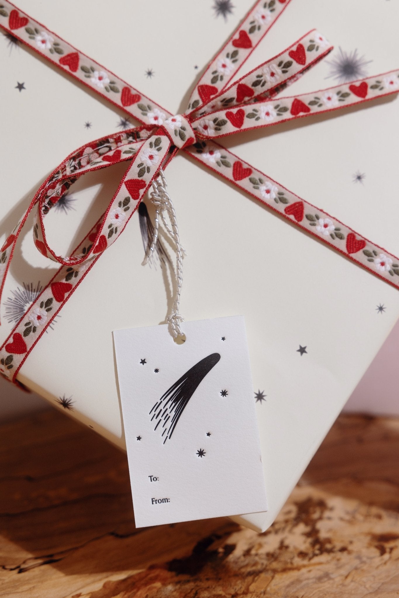 An Adelfi letterpressed gift tag with black comets, stars, and the words &quot;to&quot; and &quot;from&quot; in black ink on cream card stock attached by silver string to a present wrapped in cream and black comets Adelfi gift wrap with red hearts ribbon.