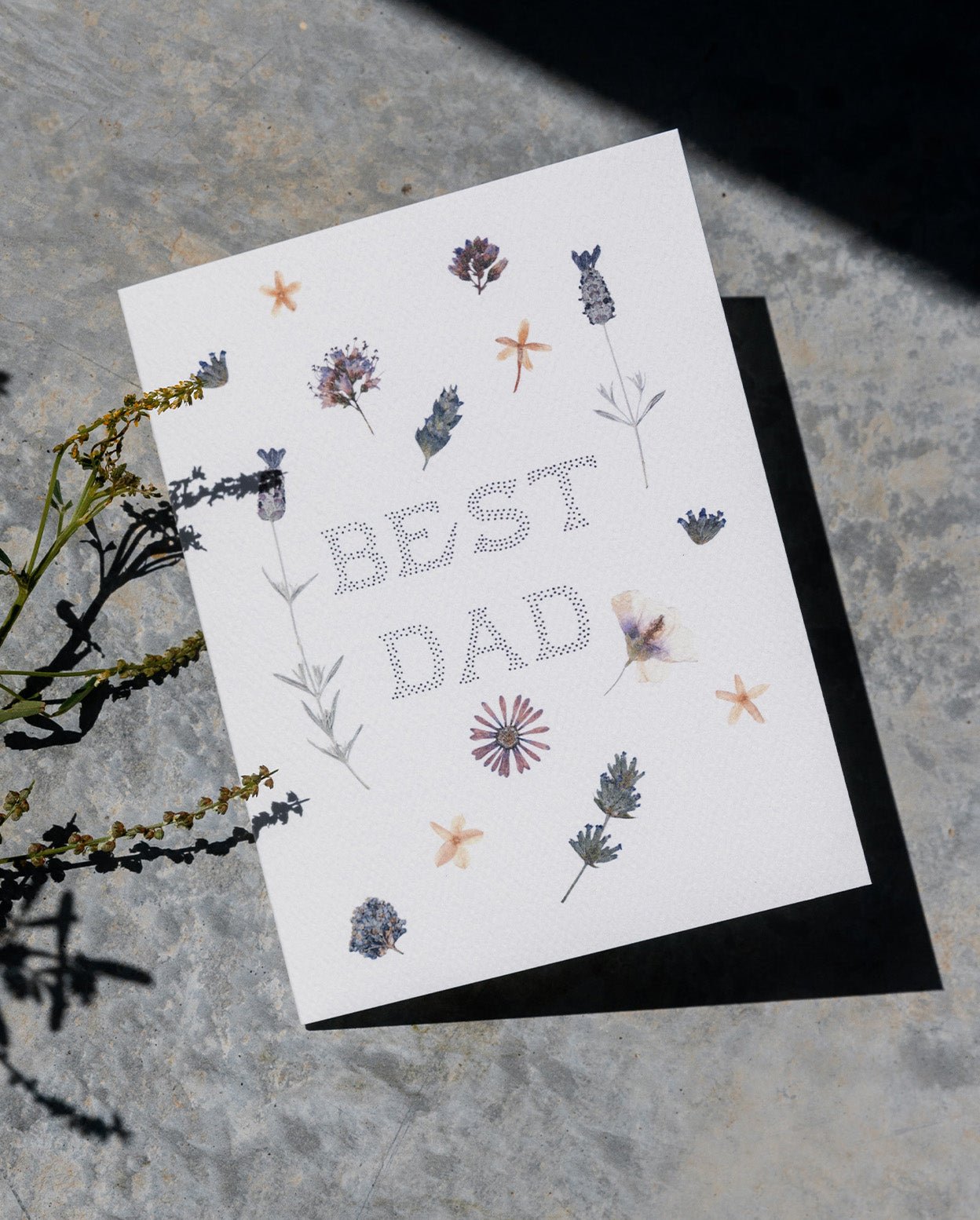 Cream colored card with scattered pressed flowers and the words &quot;Best Dad&quot; in pointillism style font. Shown on grey cement background with flowers.