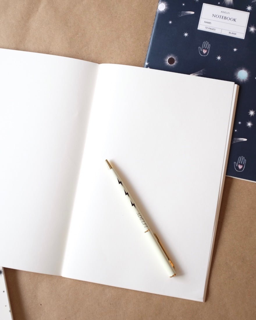 One Adelfi notebook open on a table with blank pages and an Adelfi pen. Notebook with a navy background and outer space icons in the bakground.