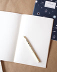One Adelfi notebook open on a table with blank pages and an Adelfi pen. Notebook with a navy background and outer space icons in the bakground.