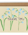 Four drooping blue forest flowers cover the card nearly top to bottom horizontally, with the words "Thank You" going through the center of them in black ink. This design is printed on a cream colored background. Shown with Kraft envelope.
