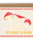 Three red mushrooms arranged above the words "my heart is mush" in orange font with red hearts stacked along every side of the card. Printed on a cream background. Shown with kraft envelope.