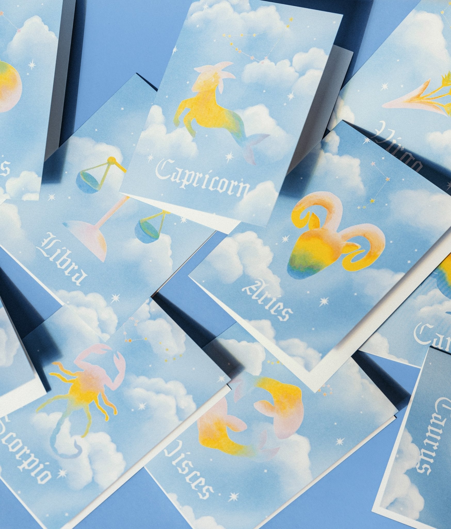 Adelfi Horoscope cards have a light blue background with white clouds and each of the twelve horoscope symbols painted in pastel rainbow colors.