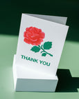 Cream colored background featuring a big red rose with a green stem and leaves, below it is written in green-colored font "Thank You" printed on cardstock on top of a white block with a green background.