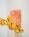 Greeting card with the word "Congrats" across the front in white hollow font with neon icons against an ombre orange background. Shown sitting on yellow orchids in a clay pot against an off white background.