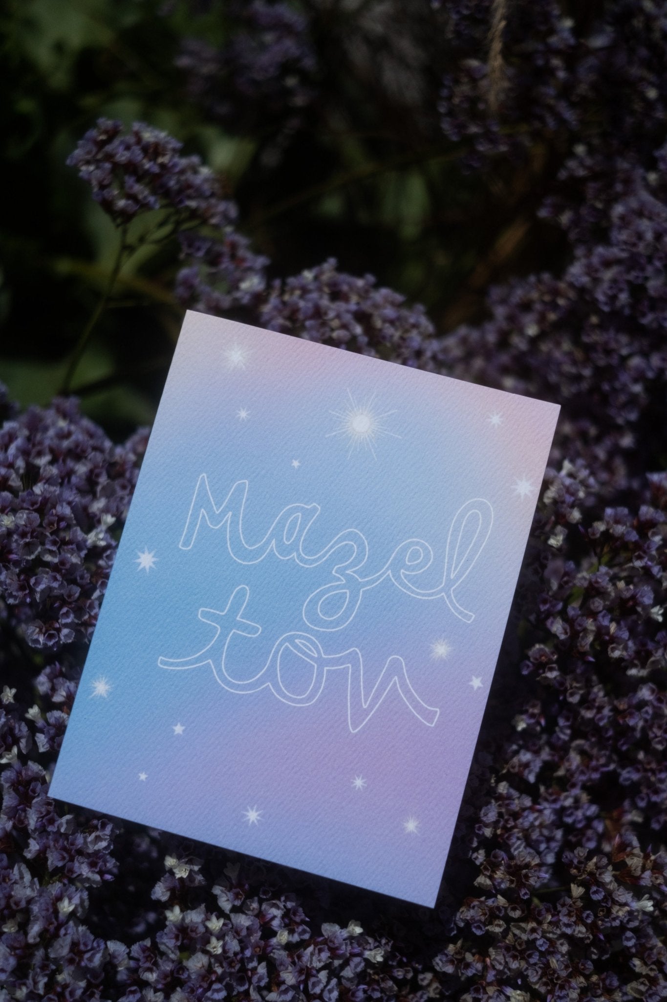 &quot;Mazel Tov&quot;  in large, hollow cursive on a gradient blue-lavender background with various kinds of stars printed on cardstock with dark purple flowers and green leaves in the background.