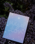 "Mazel Tov"  in large, hollow cursive on a gradient blue-lavender background with various kinds of stars printed on cardstock with dark purple flowers and green leaves in the background.
