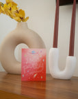 "Let's Party" printed in large cursive on an ombre red background with lightning bolts, still and shooting stars, and a blue Saturn design on a wood surface with a cream clay circular flower vase with yellow orchids on the left and a white ceramic double sided candle holder with brown candles.