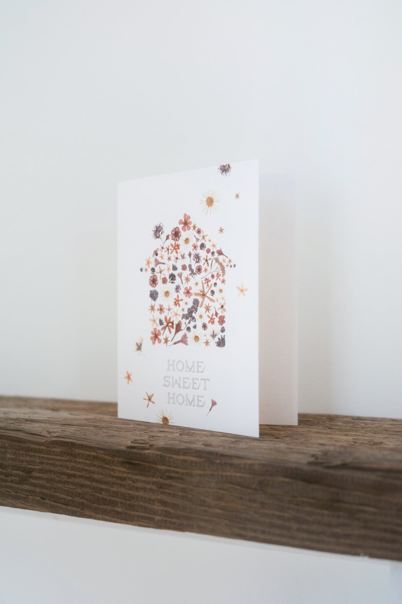 Cream colored background with pressed flowers scattered across dried flora constructed in the shape of a house with the words &quot;Home Sweet Home&quot; printed below. Shown on wood shelf with white background.