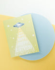 Greeting card with green background featuring stars and flying saucer with "happy birthday" printed down the front. Shown on blue platform with green and white background.