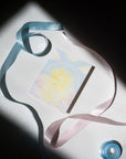 "Welcome To The World Little One" printed in white cursive font bending around a sunset over the ocean with a pink and blue cloudy landscape design printed on cardstock surrounded by pink and blue ribbon.