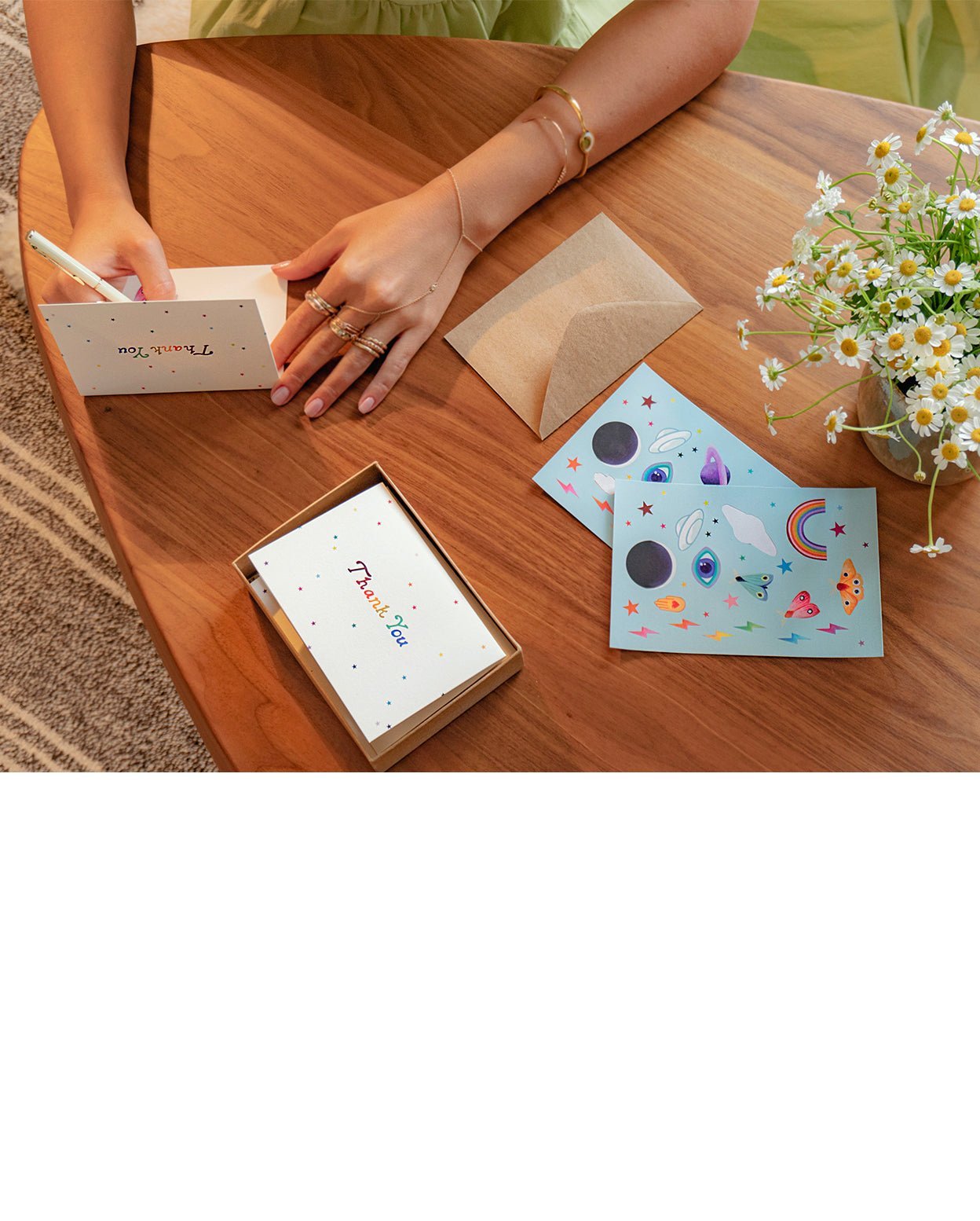 Cards modeled in use by a person with several rings and bracelets writing on the blank card on top of a wood surface with neon icons stickers next to white flowers in a clay pot.