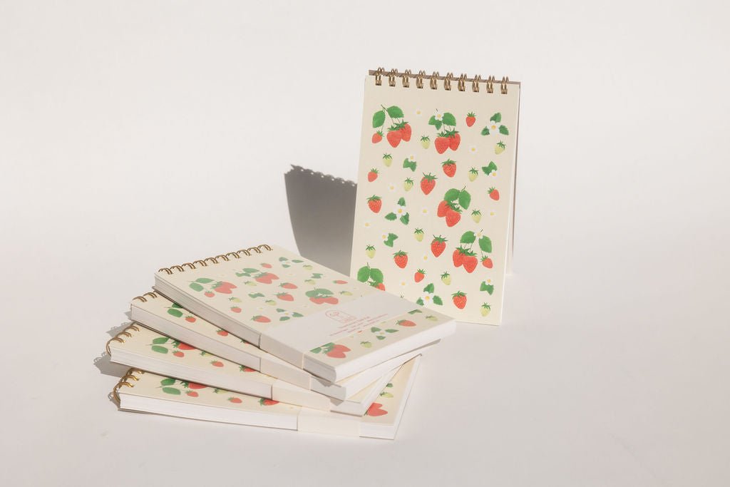 Front view of the strawberries taskpad with varying kinds of strawberries, loose and on stems, green and red, large and small, surrounded by white flowers with and without surrounding green leaves. Printed on a cream background.