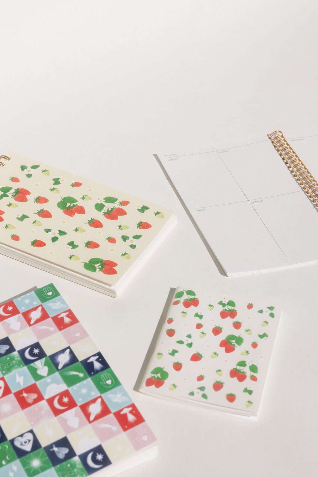 Front view of the strawberries taskpad with varying kinds of strawberries, loose and on stems, green and red, large and small, surrounded by white flowers with and without surrounding green leaves. Printed on a cream background. Shown with our planner, Strawberries pocket notebook, and Holiday Checkerboard journal.