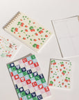 Holiday Checkerboard journal. Pink, blue, red, green, and yellow squares with neon icons of UFOs, hearts with eyes, moths, moons with stars, Saturn, shooting stars, moon phases, lightning bolts, moths, Hamsa hands, and a brightly burning star. Shown with our Strawberries & Cream taskpad, Strawberries pocket notebook, and planner.