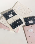 Adelfi notebook trio on a white background. One notebook has a cream background with black stars and comets, the second is navy blue with mystical outer space icons, and the third is pink with stars and eclipsing moons.