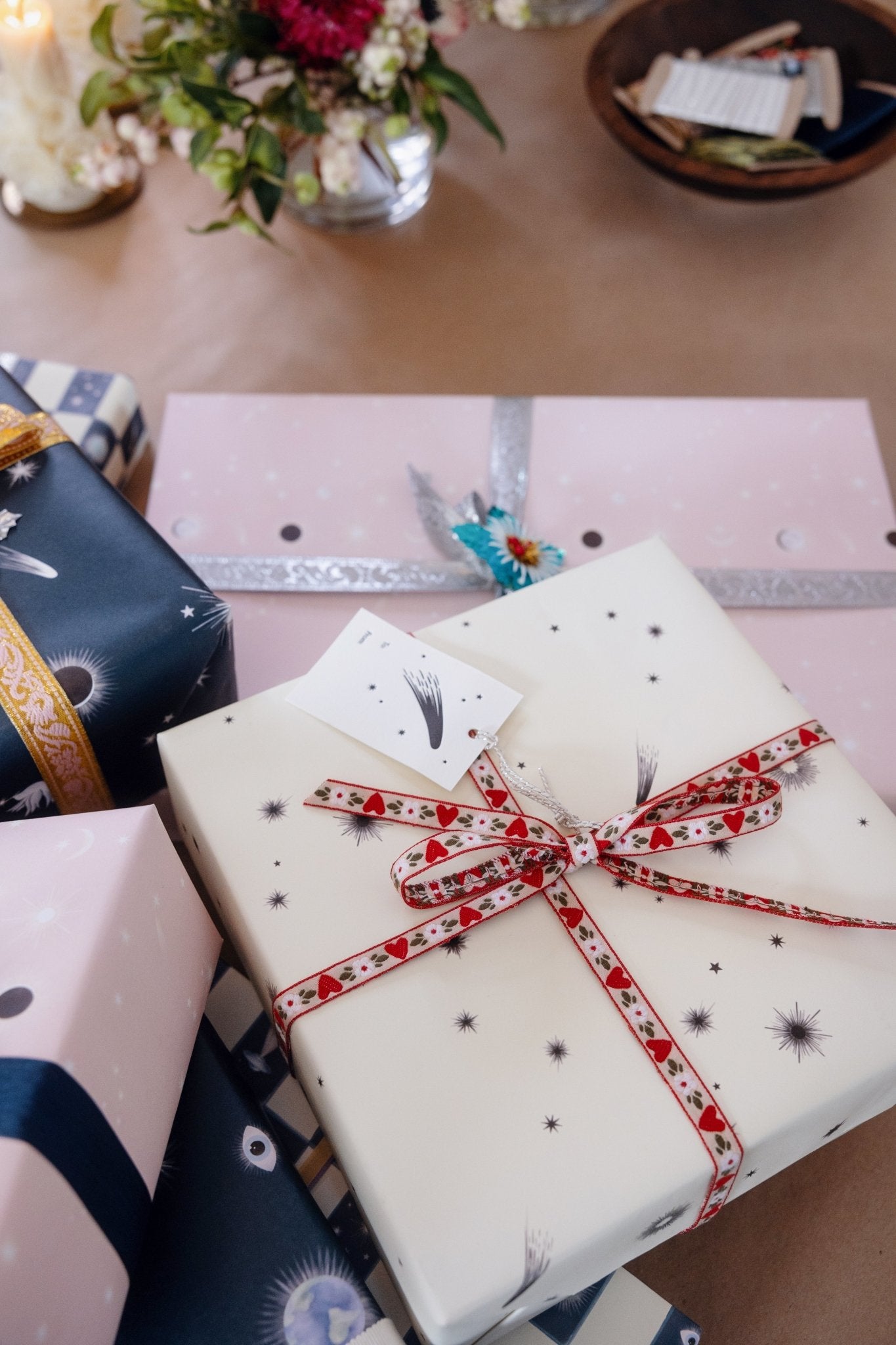 A pile of presents wrapped in assorted Adelfi gift wrap and ribbons on a table covered in kraft paper with flowers and a bowl of ribbons in the background.