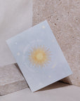 Greeting card with a place blue background, stars, and the words "Another Trip Around the Sun" circling around a yellow sun. Card is propped up on a beige background and is leaning at an angle against a light gray stone.