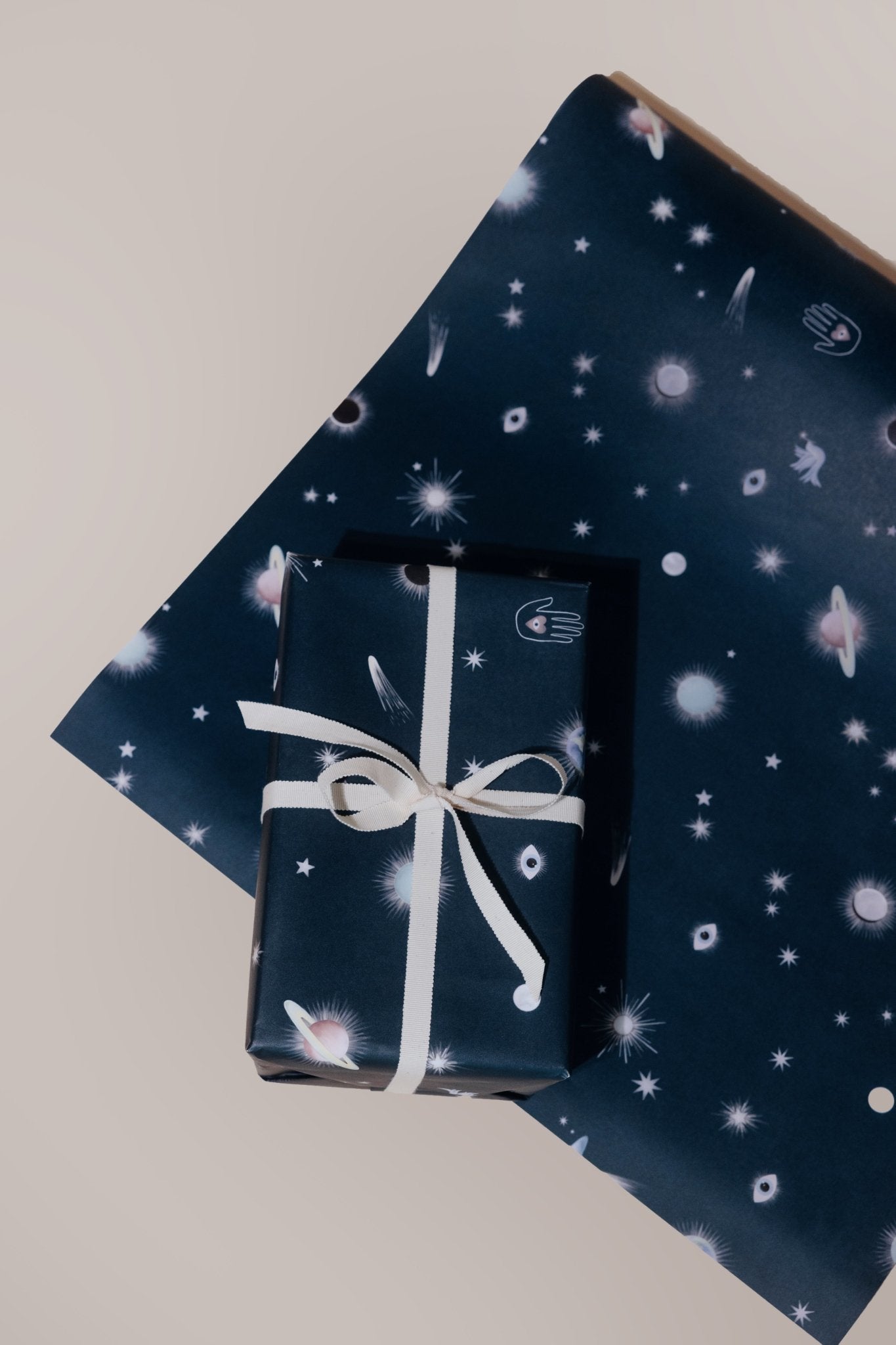 A present with a white box wrapped in Adelfi "Galaxy" gift wrap, dark blue background with scattered with UFOs, shooting stars, evil eyes, planets, doves, and Hamsa hands, on a sheet on of "Galaxy" gift wrap.
