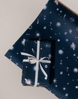 A present with a white box wrapped in Adelfi "Galaxy" gift wrap, dark blue background with scattered with UFOs, shooting stars, evil eyes, planets, doves, and Hamsa hands, on a sheet on of "Galaxy" gift wrap.