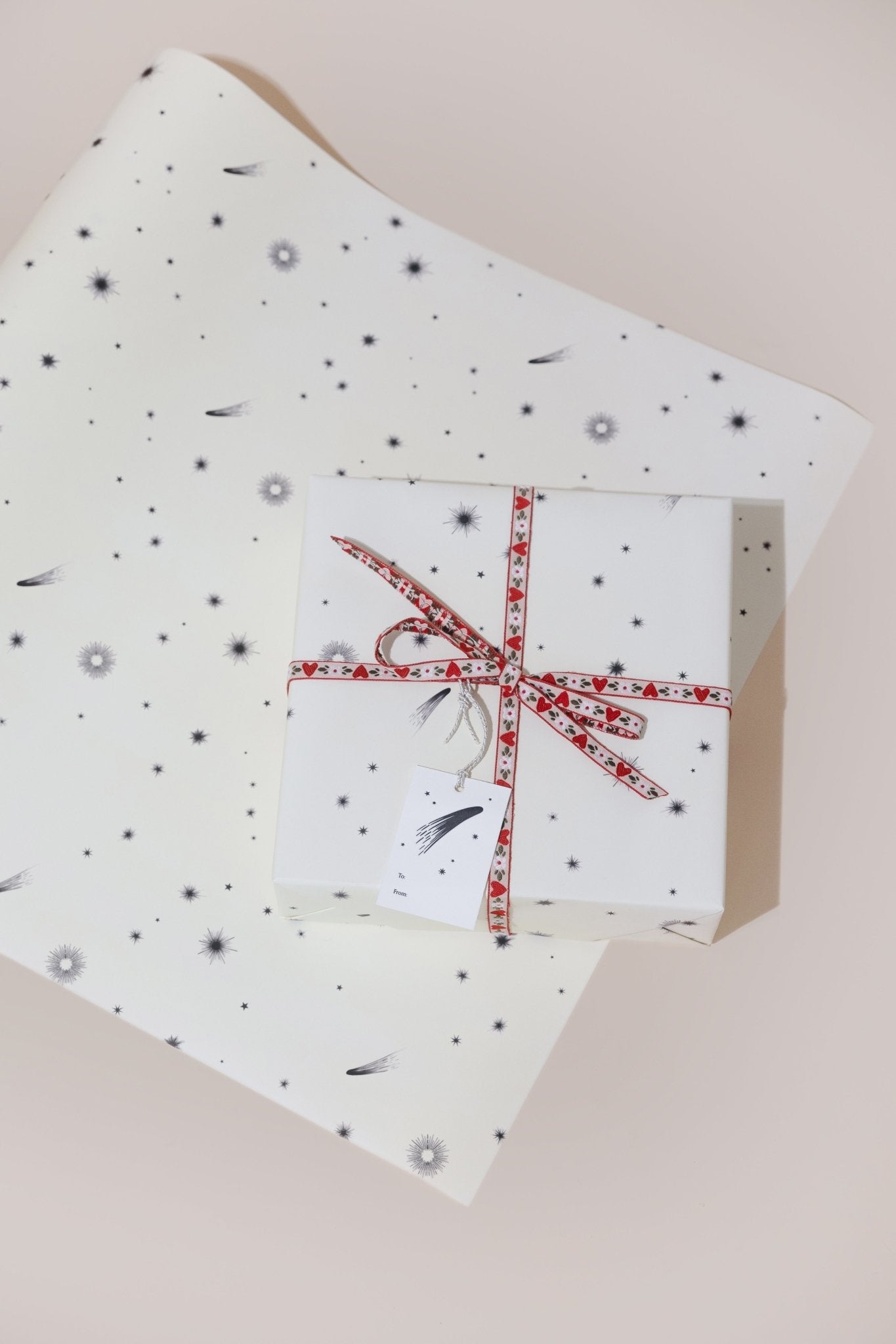 A present wrapped in gift wrap with a cream background scattered with black comets and stars and a red bow, placed on a single flat sheet of the same gift wrap.