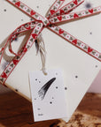 An Adelfi letterpressed gift tag with black comets, stars, and the words "to" and "from" in black ink on cream card stock attached by silver string to a present wrapped in cream and black comets Adelfi gift wrap with red hearts ribbon.