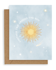 Greeting card with a pale blue background and scattered stars, a big yellow sun in the middle is circled by the words "Another Trip Around the Sun." Shown with kraft envelope.