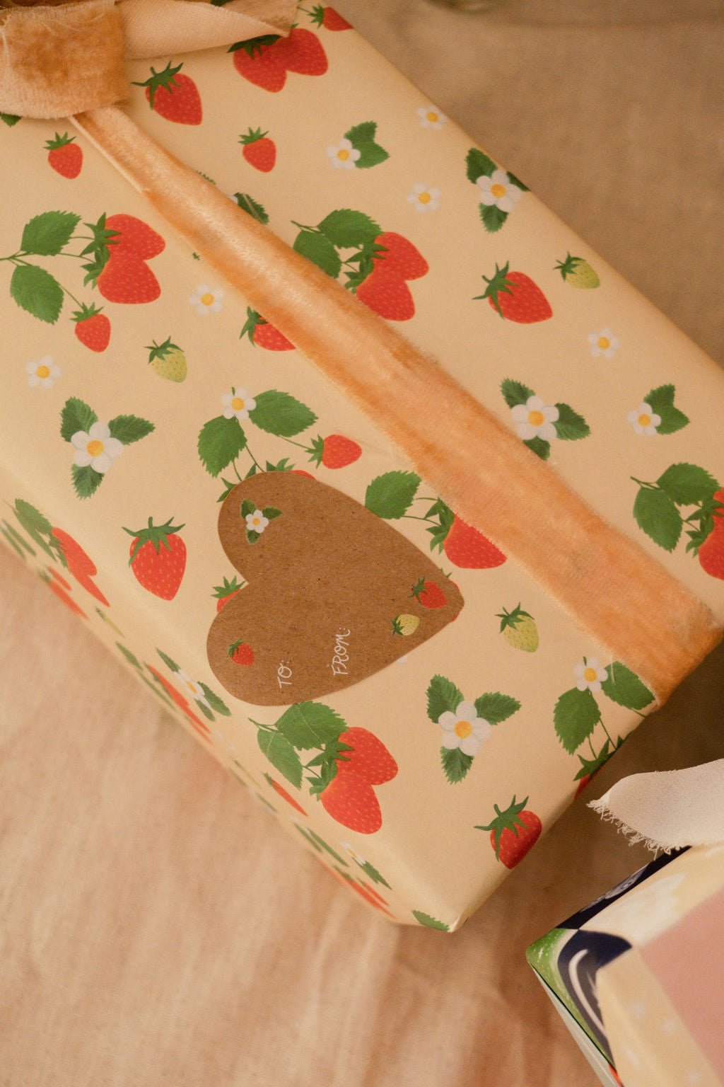 This Gift Sticker is heart-shaped with a red strawberry in the upper left hand corner, a green and red strawberry at the bottom, and a white flower with three leaves in the upper right corner, with the words &quot;To&quot; and &quot;From&quot; written in cursive in the center left. Shown on our Strawberries gift wrap.
