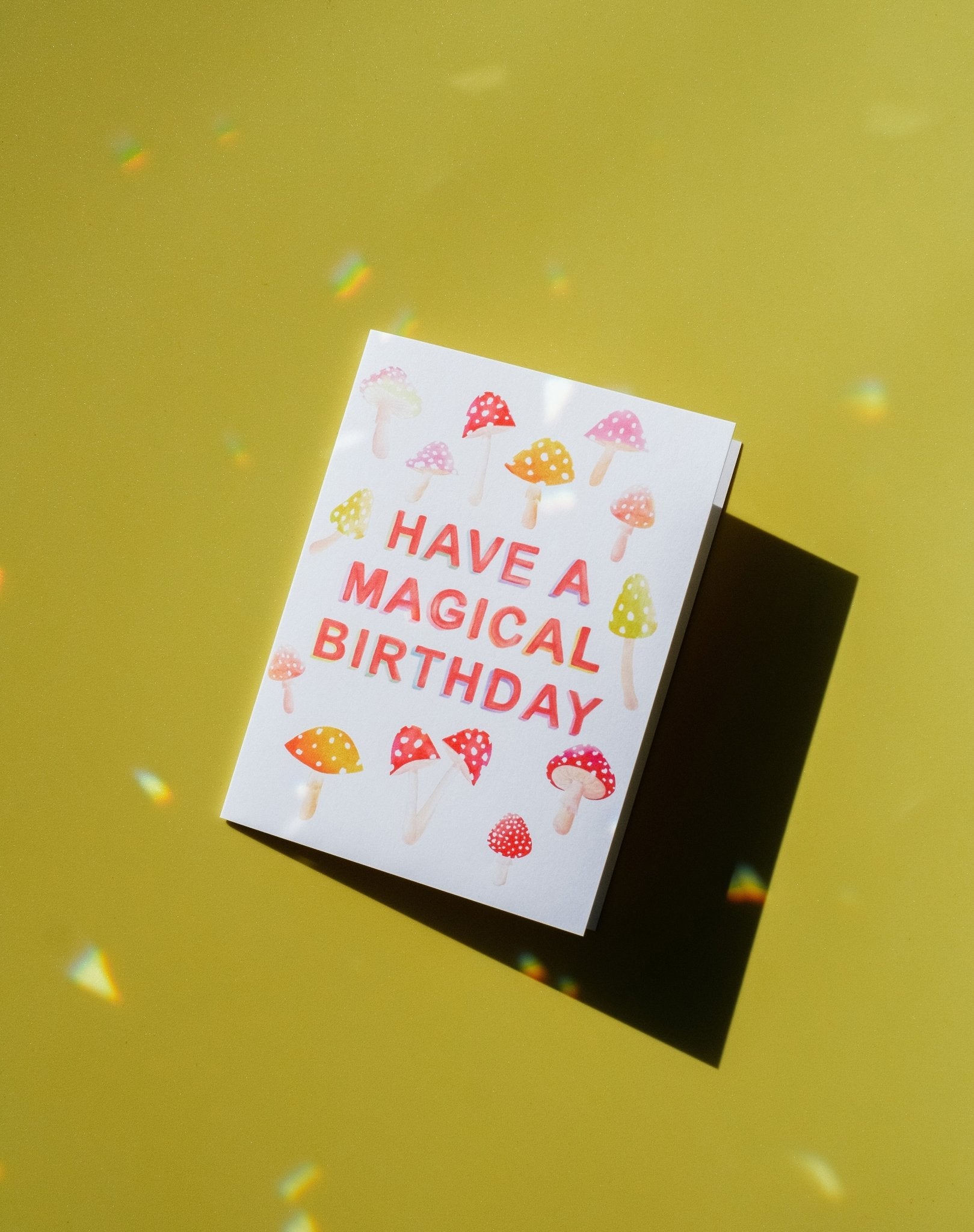 Cream colored card with pink, orange and red mushrooms and red printed text with the words "Have A Magical Birthday" in the middle. Shown on green background.