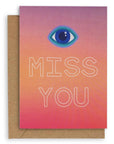 Greeting card with the symbol of an eye followed by the words "Miss You" in white hollow font on a gradient purple, orange and red background. Shown with brown kraft paper envelope.