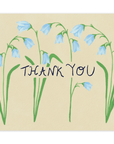 Four stems of drooping blue forest flowers cover the card nearly top to bottom horizontally, with the words "Thank You" going through the center of them in black ink. This design is printed on a cream colored background. 