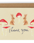 Our new Forest Creatures design! This card features two light brown/dirty blonde rabbits facing each other with a red magic mushroom between them and two red mushrooms on either side of the words "Thank You" printed in black ink on a cream background. Shown with Kraft envelope.