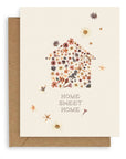 Cream colored background with pressed flowers scattered across dried flora constructed in the shape of a house with the words "Home Sweet Home" printed below. Shown with Kraft envelope.