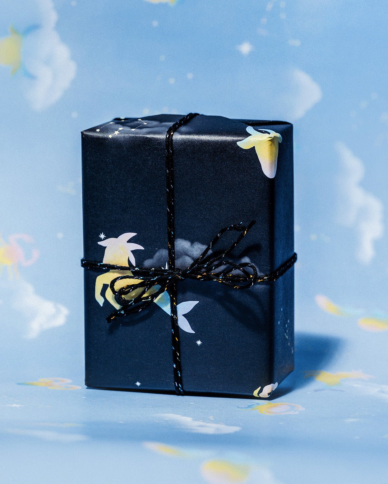 Black horoscope gift wrap tied with black and white string against a blue sky background with clouds.