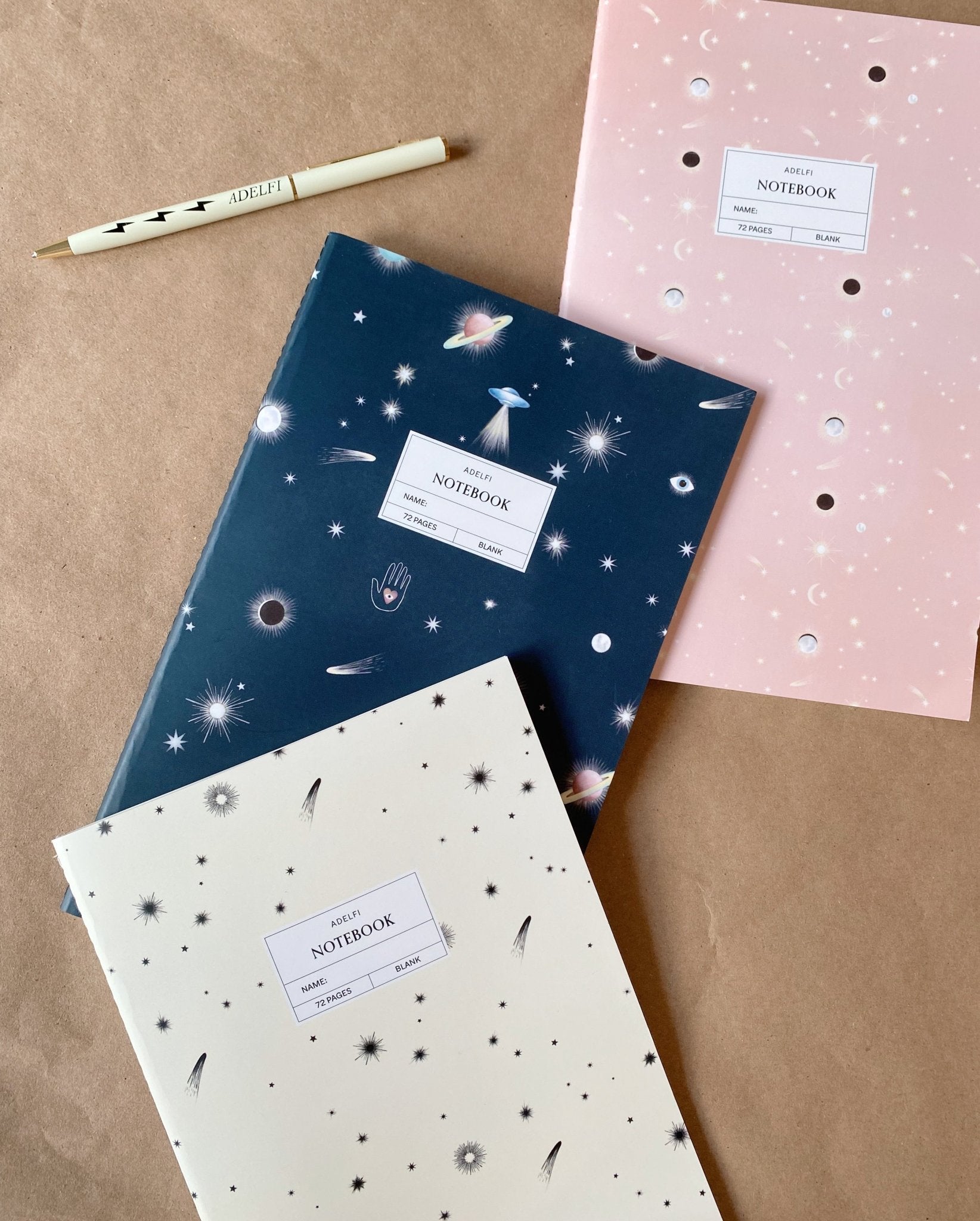 Adelfi notebook trio on a kraft paper background with an Adelfi pen. One notebook has a cream background with black stars and comets, the second is navy blue with mystical outer space icons, and the third is pink with stars and eclipsing moons.