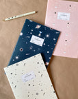 Adelfi notebook trio on a kraft paper background with an Adelfi pen. One notebook has a cream background with black stars and comets, the second is navy blue with mystical outer space icons, and the third is pink with stars and eclipsing moons.