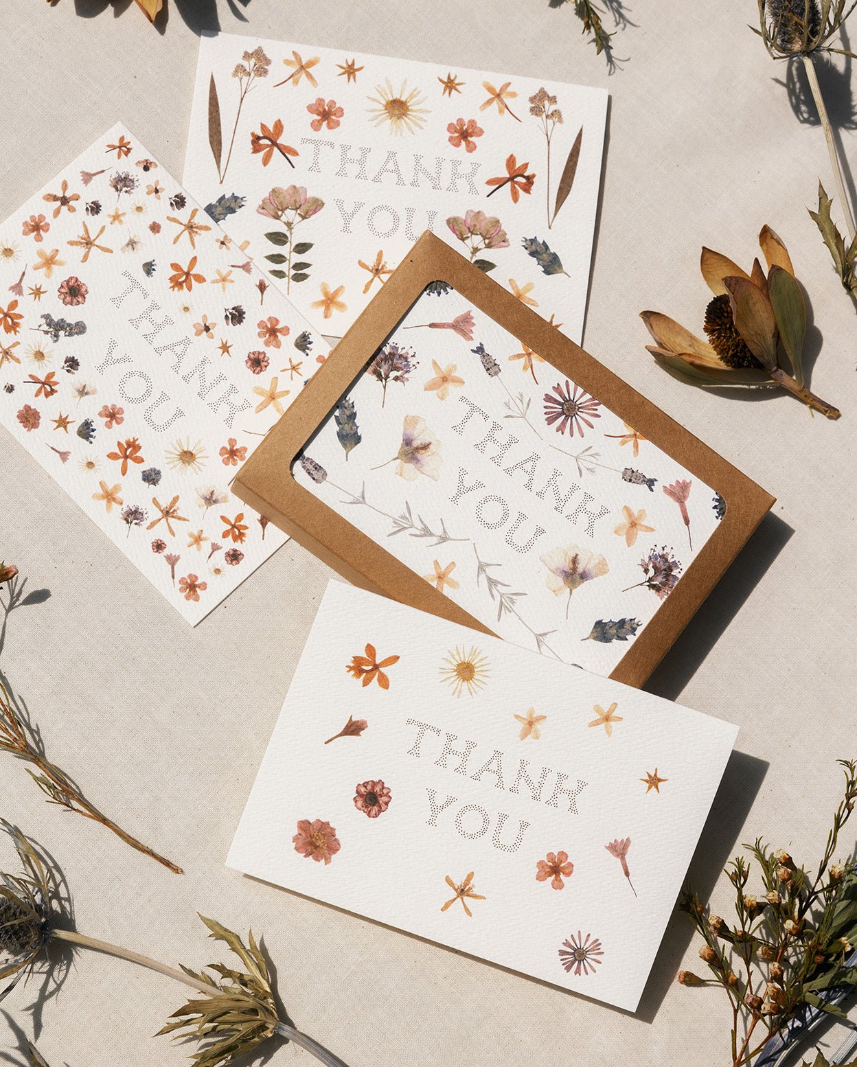 Four Adelfi thank you cards featuring designs using pressed flowers and the words &#39;Thank You&#39; in the center. Cards are placed on white canvas background with various flora.
