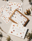 Four Adelfi thank you cards featuring designs using pressed flowers and the words 'Thank You' in the center. Cards are placed on white canvas background with various flora.
