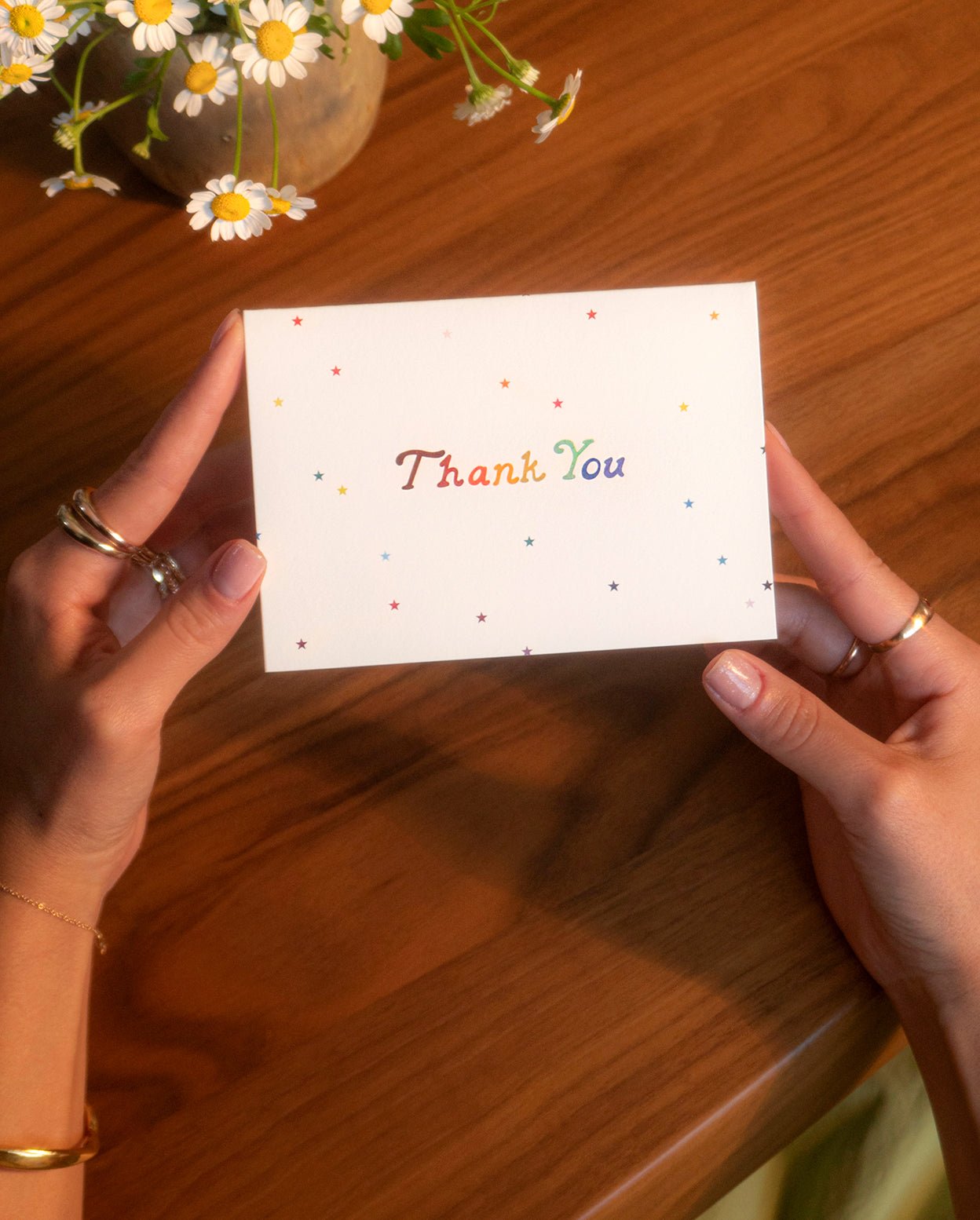  Rainbow stars thank you card with rainbow colored letters printed on white cardstock. Hands modeling card above a wood surface with white flowers in a clay pot in the background.